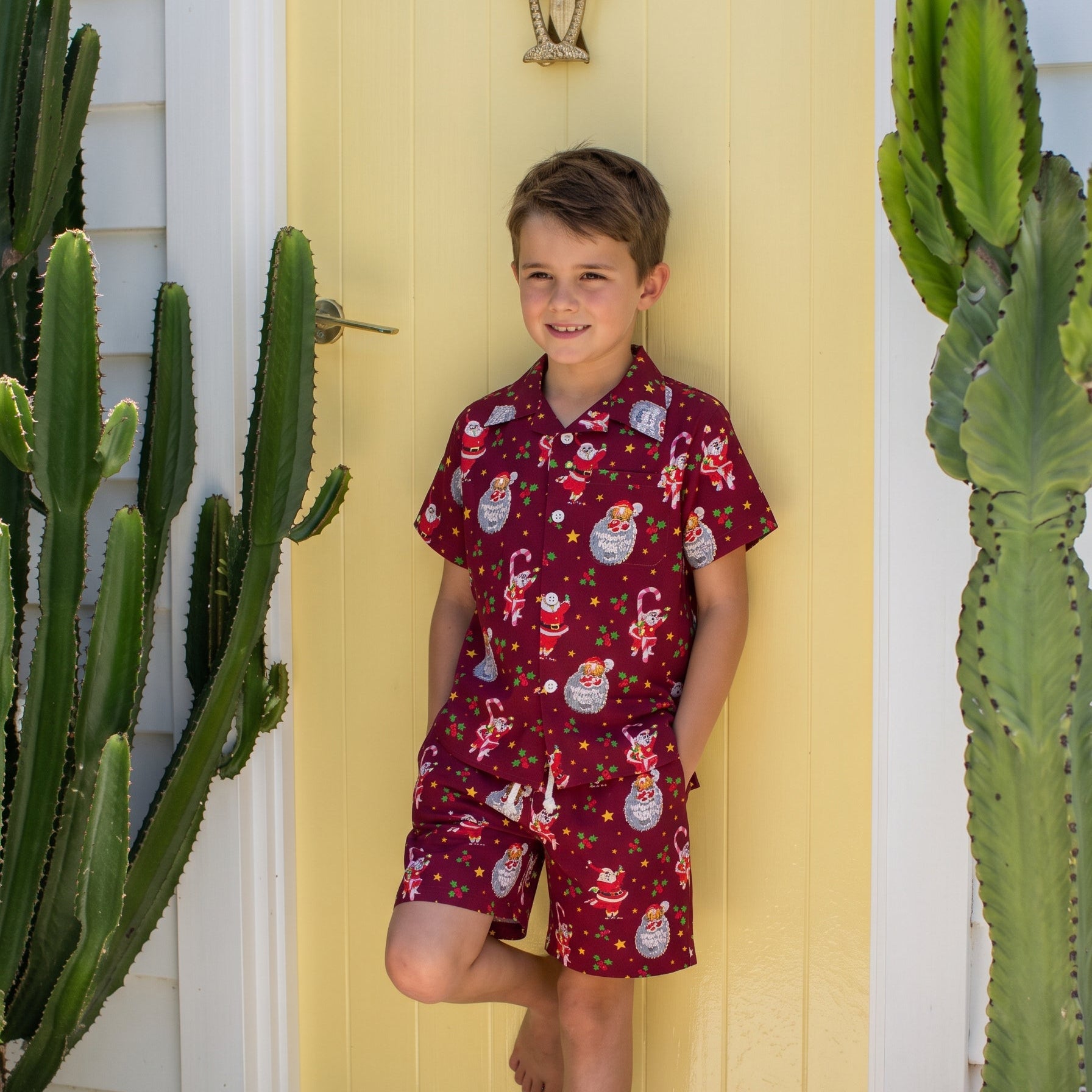 A young boy stands barefoot in front of a yellow door, wearing a matching short-sleeve pajama set with a red base and a festive Santa Claus snowman print. The scene is framed by tall potted cacti on both sides.