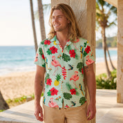 A man wearing a light mint green short-sleeve button-up shirt with a colorful tropical print of flamingos, palm leaves, and parrots, standing on a beachside walkway.