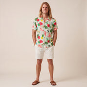 A man stands against a plain beige studio background wearing a short-sleeve tropical print button-up shirt with white shorts and brown sandals. The shirt features a light mint green base with pink flamingos, red hibiscus flowers, green leaves, and yellow pineapples.