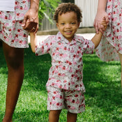 A family of three wearing matching floral-print loungewear sets stands on a grassy lawn outdoors. The set includes a short-sleeve button-up shirt and shorts for the adults, and a matching tunic dress and shorts set for the child.