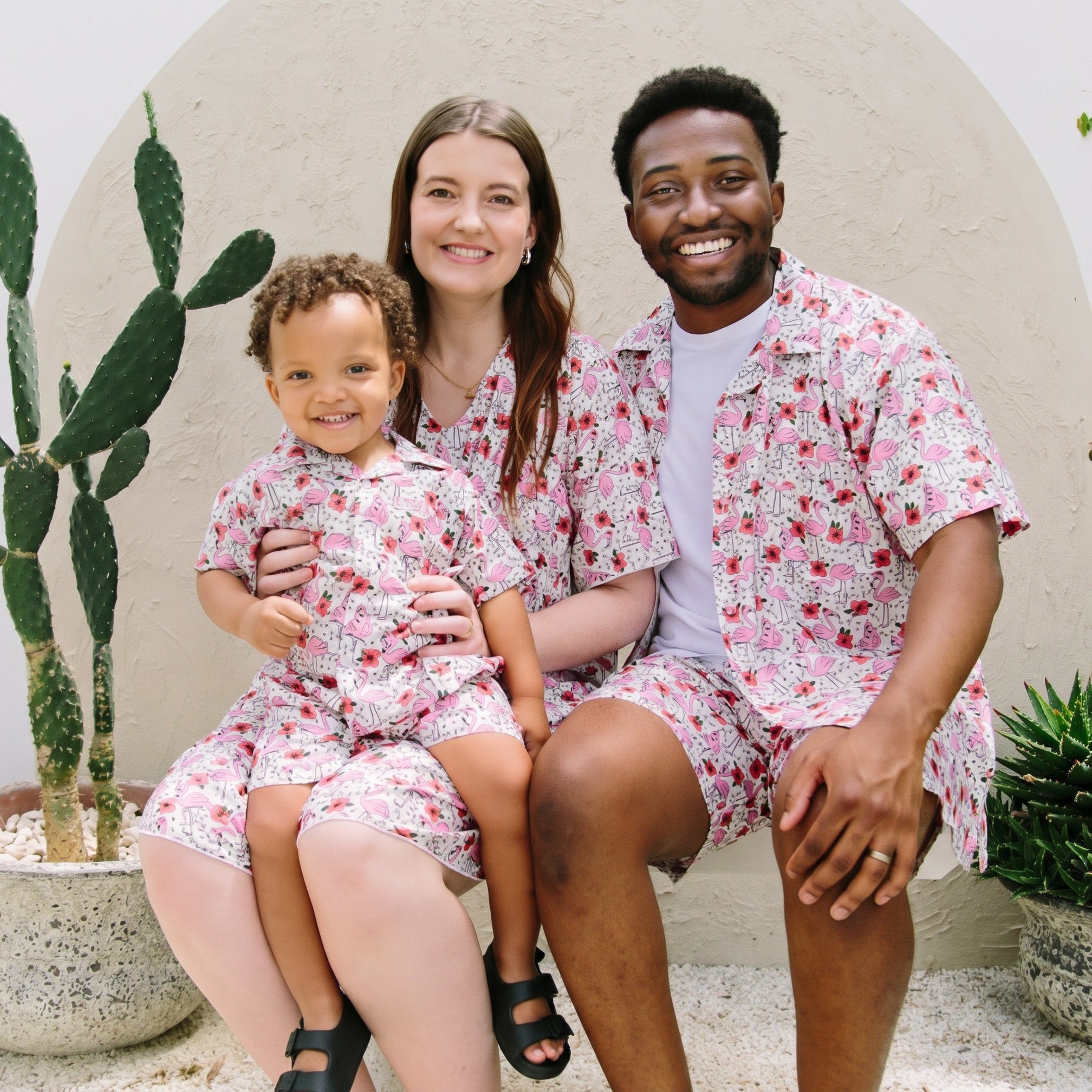 A family of three wearing matching short-sleeve button-up shirts and shorts sets with a pink and red strawberry print on a white background, posed together in front of a stucco wall with potted cacti.