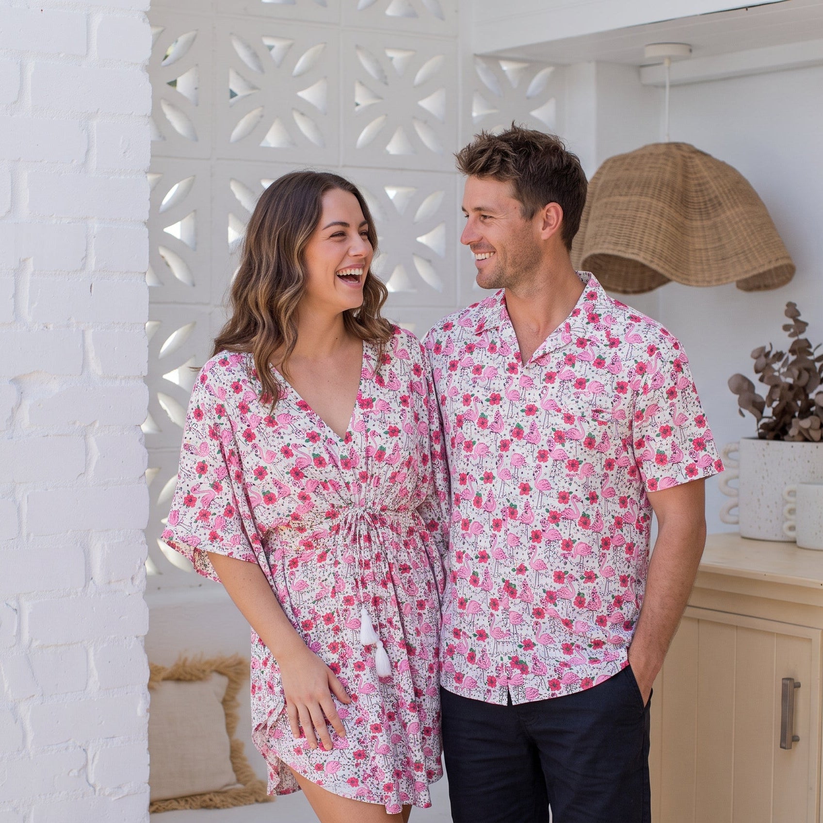 A man and a woman standing together in a bright, modern room, wearing matching pink and white floral short-sleeve button-up shirts and matching floral tie-dye shorts.