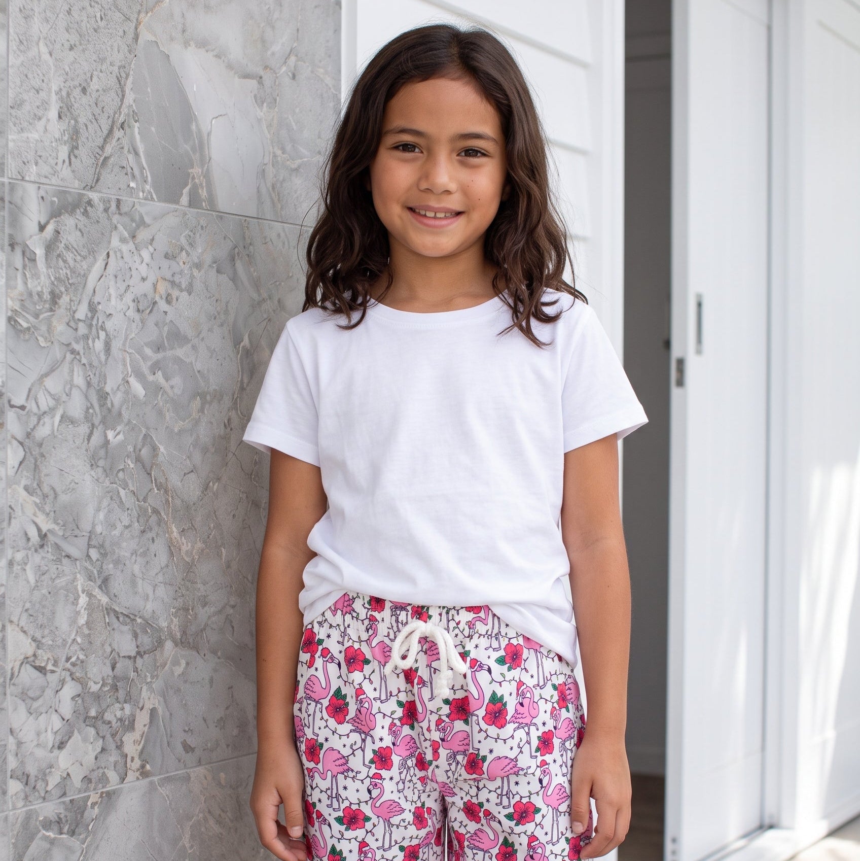 A young girl stands outdoors wearing a white t-shirt and pink floral print shorts with a white drawstring, posing in front of a gray marble wall and a white sliding door.