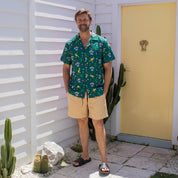 A man stands outdoors in front of a white house wall and a yellow door, wearing a green short-sleeve button-up shirt with a colorful dinosaur and skull print, tan shorts, and black slide sandals.