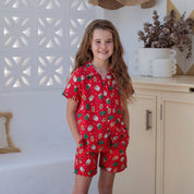 A young girl stands in a bright, modern room wearing a red short-sleeve matching set with a white strawberry pattern. The set includes a short-sleeve collared shirt and matching shorts.