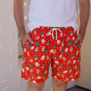 A man wearing red patterned swim trunks with a white drawstring, a plain white t-shirt, and black sunglasses, standing in a bright, modern indoor setting.