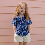 A young girl with long red hair stands barefoot on a green lawn in front of a pink house siding, wearing a blue short-sleeve button-up shirt with a white tree and snow pattern, paired with cream shorts.