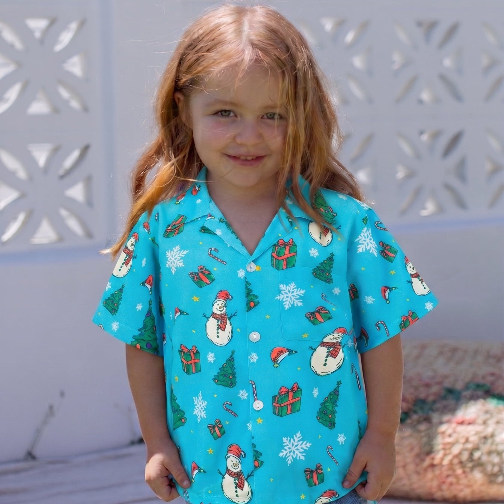 A young girl with long light brown hair stands outdoors wearing a bright turquoise short-sleeve button-up shirt with a festive snowman and Christmas print, paired with blue jeans.