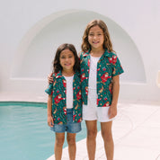 Two young girls standing barefoot by a swimming pool, wearing matching green short-sleeve button-up shirts with a red and yellow holiday print, white t-shirts, and white shorts.