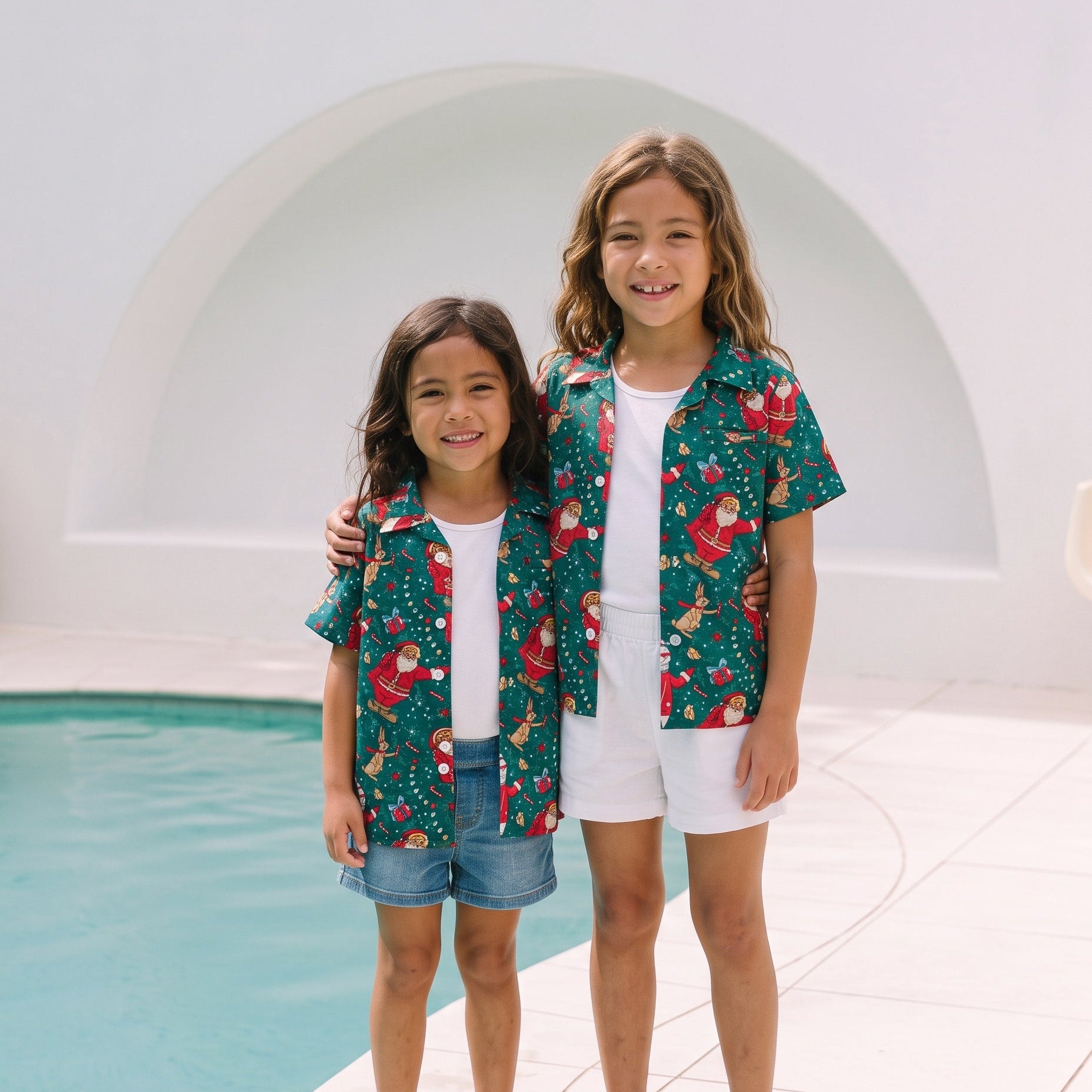 Two young girls standing barefoot by a swimming pool, wearing matching green short-sleeve button-up shirts with a red and yellow holiday print, white t-shirts, and white shorts.