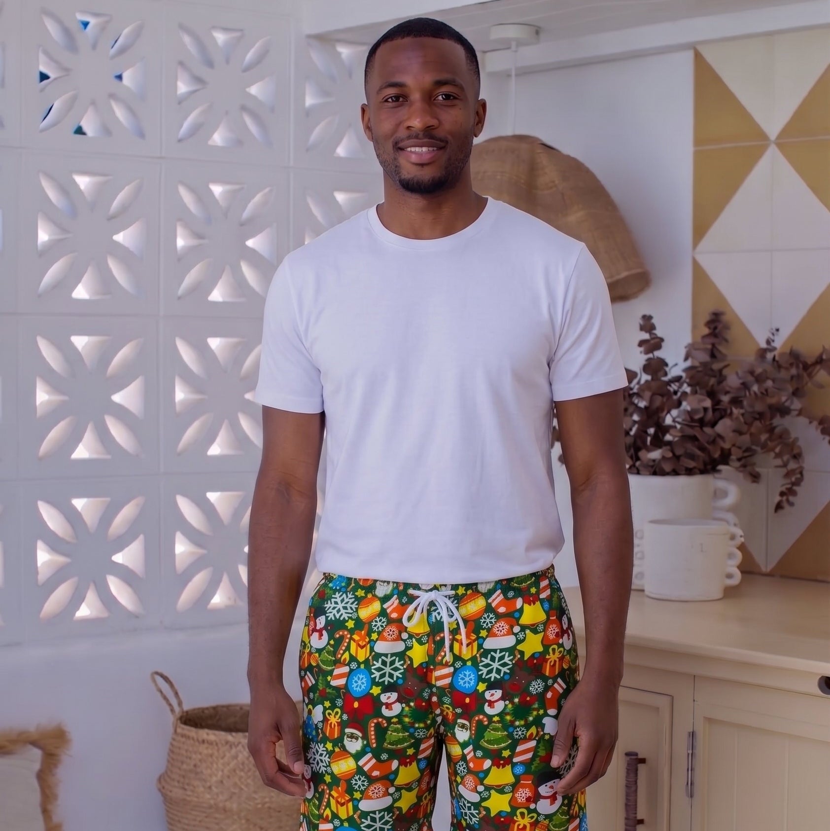 A man wearing a white t-shirt and colorful patterned swim trunks stands in a bright, modern kitchen setting.