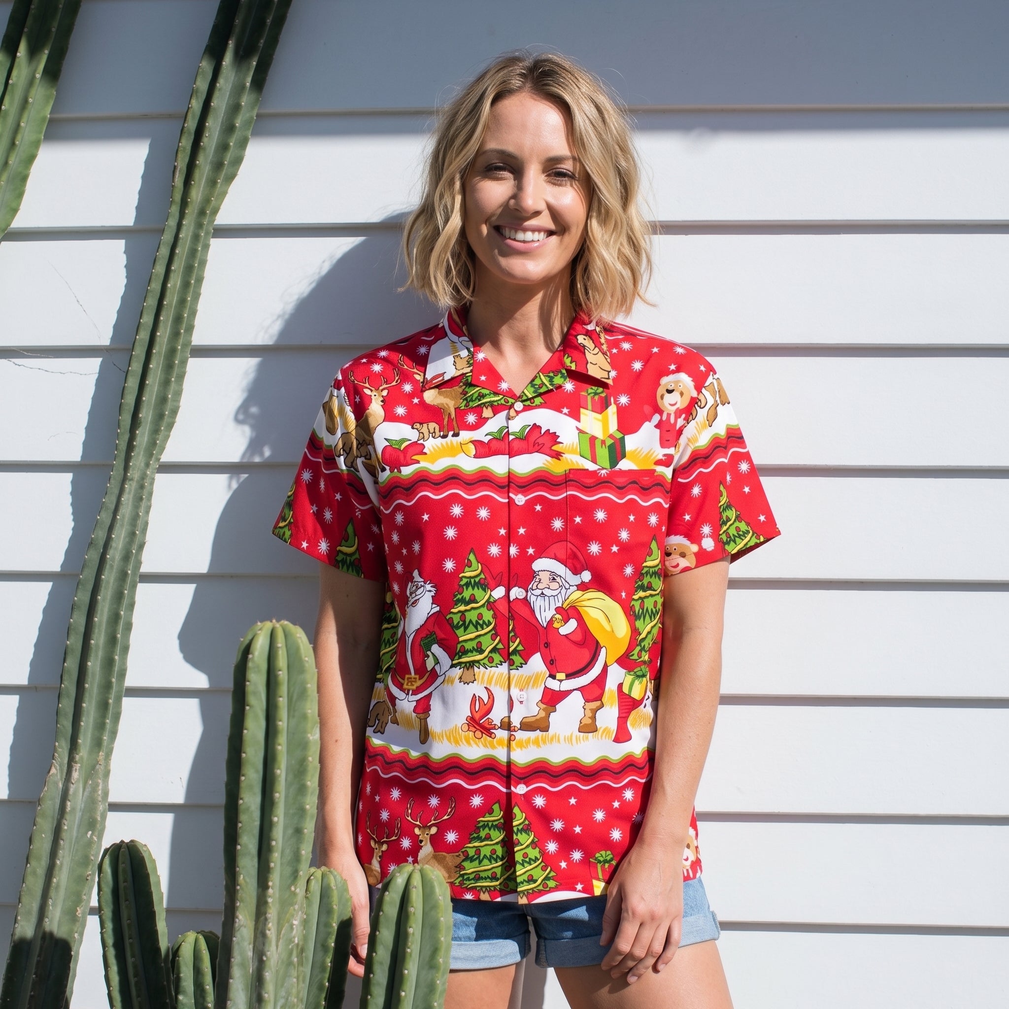 A woman stands outdoors against a white siding wall wearing a bright red short-sleeve button-up shirt with a festive Christmas print featuring Santa Claus, a dog, trees, and snowflakes. She is also wearing denim shorts, with tall cacti visible on the left side of the frame.