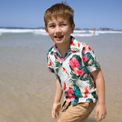 A young boy smiling on a sunny beach, wearing a white short-sleeve Hawaiian-style button-up shirt with a vibrant green, pink, and orange tropical floral print, paired with tan pants.