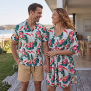 A man and a woman standing on a wooden deck wearing matching tropical floral print swim shirts and a matching women's dress. The print features pink hibiscus flowers, green leaves, and orange koi fish on a white background.