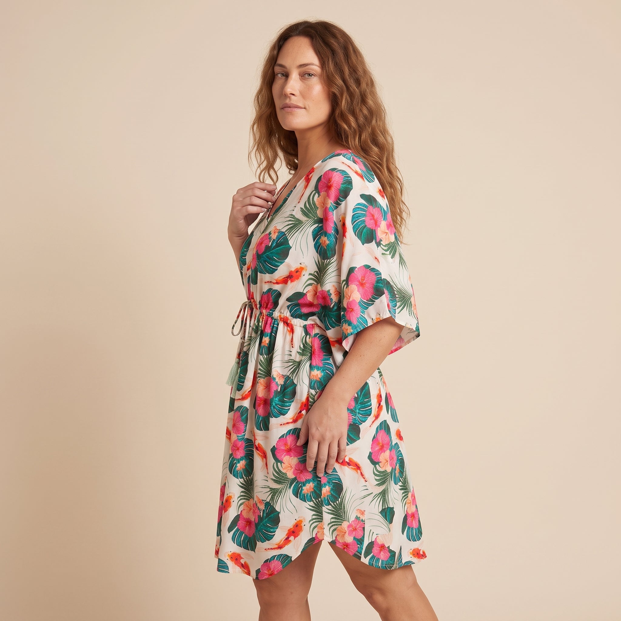 A woman modeling a short tropical print dress with pink hibiscus flowers and green palm leaves on a cream background, set against a solid beige studio backdrop.