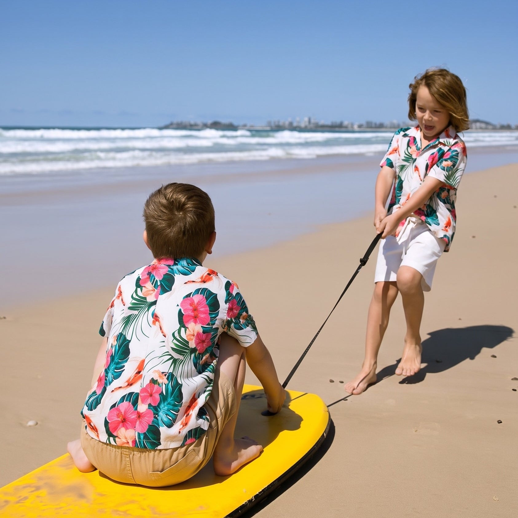Two children in matching tropical floral shirts and white shorts on a sandy beach, with one sitting on a yellow board and the other standing and holding a leash.