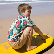 A young boy sitting on a yellow surfboard on a sandy beach, wearing a green tropical floral short-sleeve shirt and khaki shorts, with the ocean and blue sky in the background.