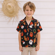 A young boy stands against a white wooden wall wearing a black short-sleeve button-up shirt with a colorful Christmas-themed print, paired with khaki shorts.