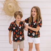 Two children standing outdoors against a white paneled wall, wearing matching black short-sleeve button-up shirts with a colorful holiday print. The boy wears khaki shorts and sandals, and the girl wears white shorts.