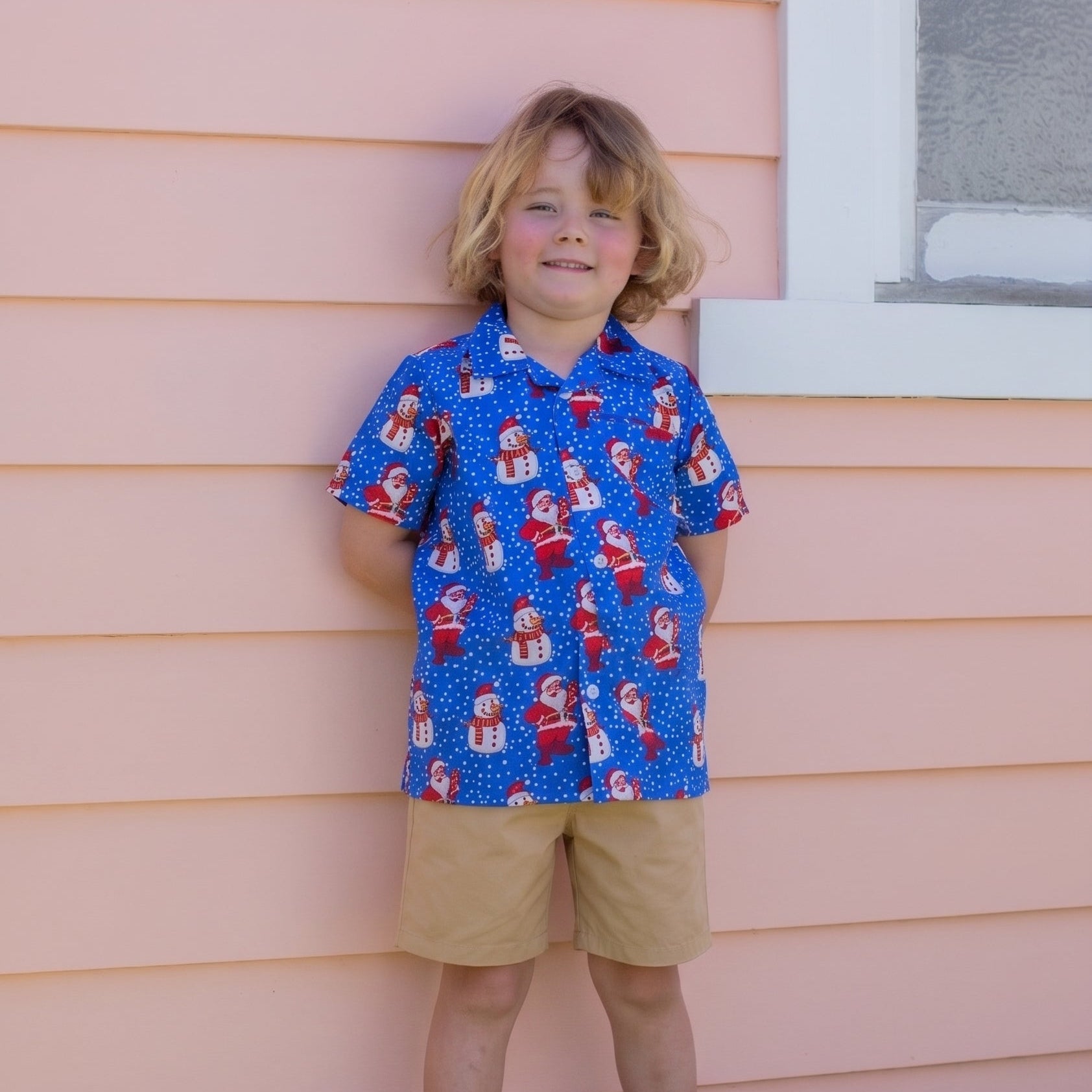 A young boy with blonde hair stands against a peach-colored house siding, wearing a blue short-sleeve button-up shirt with a red and white Santa Claus pattern, khaki shorts, and white sneakers.