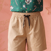 A person wearing beige drawstring shorts and a green holiday-print top, standing against a pink wall with decorative gold line art.