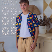 A young man wearing a blue short-sleeve button-up shirt with a colorful burger print, a white t-shirt, and beige shorts, standing in a bright, modern room with white walls and decorative panels.