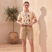 A woman stands in a warm, sunlit studio setting wearing a short-sleeve collared shirt with a colorful animal print and beige shorts with a black drawstring. She is posed against a textured beige wall with a large shadow, with a potted olive tree in the background.