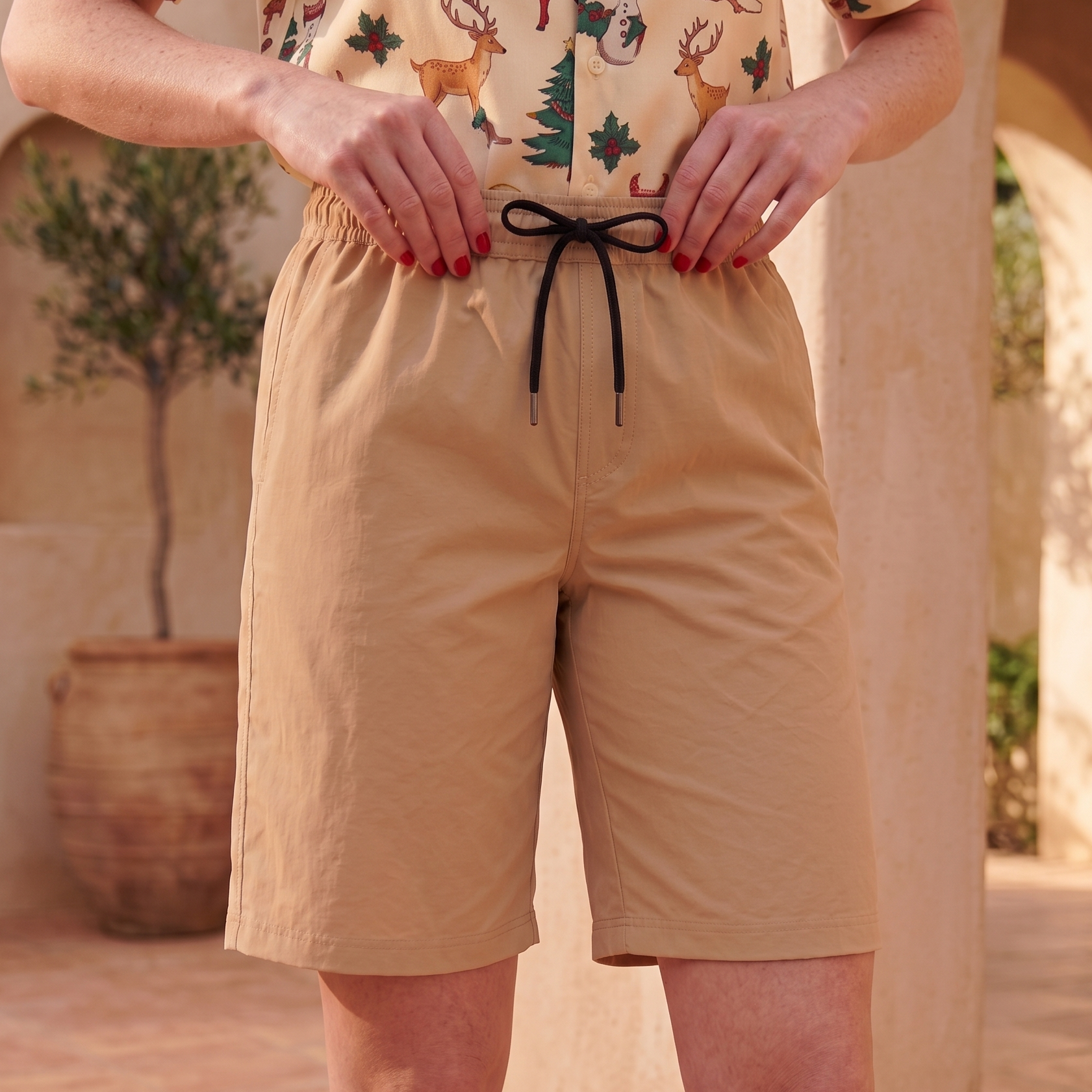 A person wearing tan drawstring shorts and a festive Christmas print shirt, standing in a sunlit outdoor patio setting with stone columns and a potted tree.