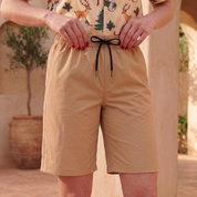 A person wearing tan drawstring shorts and a festive Christmas print shirt, standing in a sunlit outdoor patio setting with stone columns and a potted tree.