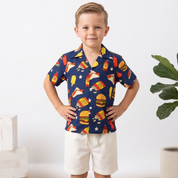 A young boy stands with hands on hips wearing a navy blue short-sleeve button-up shirt with a colorful fast-food print, paired with cream shorts and white sneakers, in a bright indoor setting.