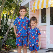 Two children standing outdoors in matching blue short-sleeve pajama sets with a red and yellow Santa Claus print, posed in front of a pink house with a yellow-and-white awning.