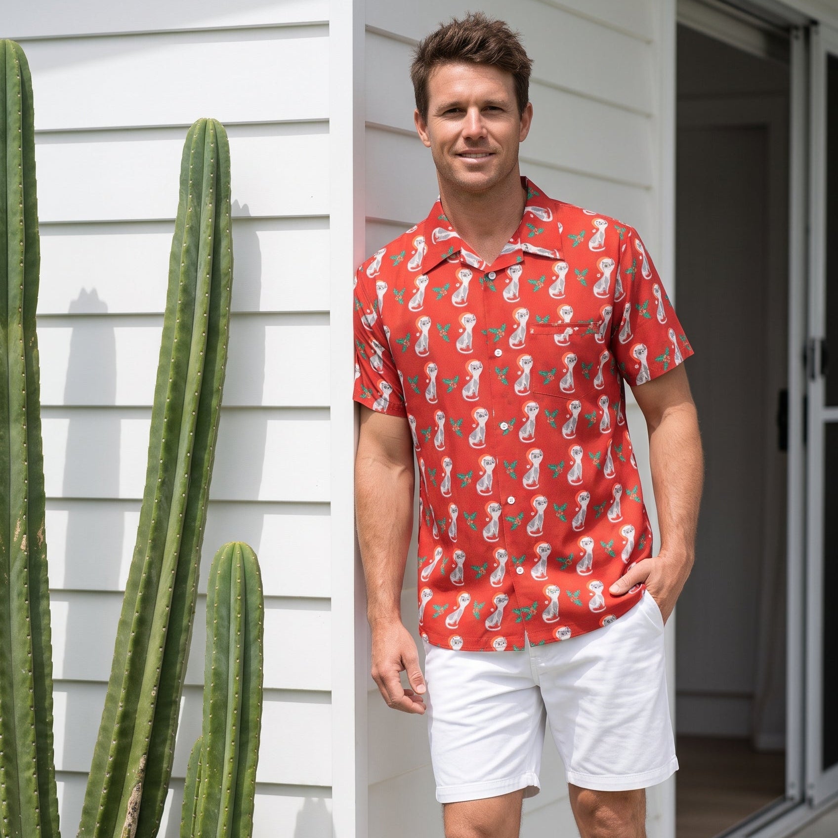 A man wearing a red short-sleeve button-up shirt with a white seahorse pattern, white shorts, and tan slide sandals, standing on a white porch next to a tall green cactus.