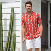 A man wearing a red short-sleeve button-up shirt with a white seahorse pattern, white shorts, and tan slide sandals, standing on a white porch next to a tall green cactus.