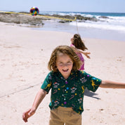 A young boy with curly brown hair runs and smiles on a sandy beach, wearing a dark green short-sleeve button-up shirt with a colorful candy cane and space-themed print, paired with khaki pants. Other children and a rocky shoreline are visible in the background.