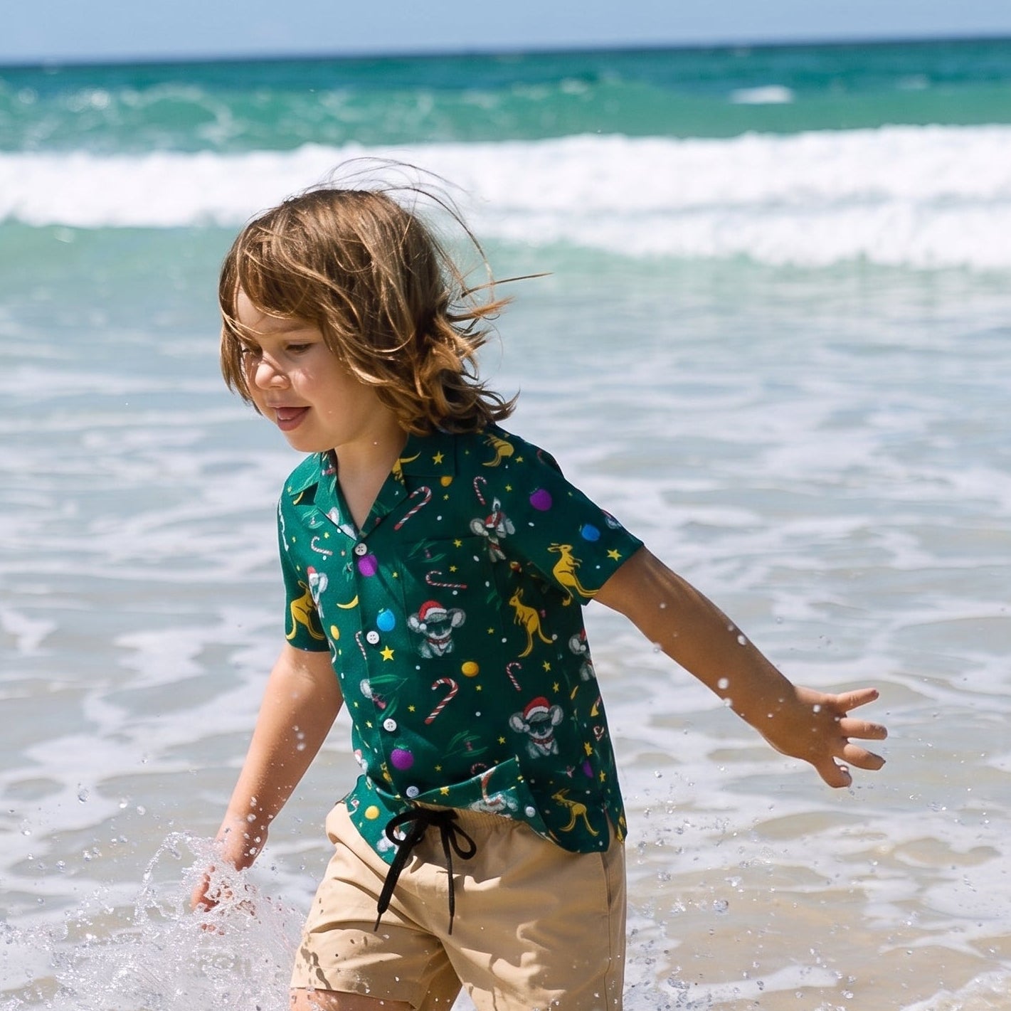 A young boy with light brown hair runs through splashing ocean water on a beach, wearing a dark green short-sleeve button-up shirt with a colorful cartoon print and beige shorts.