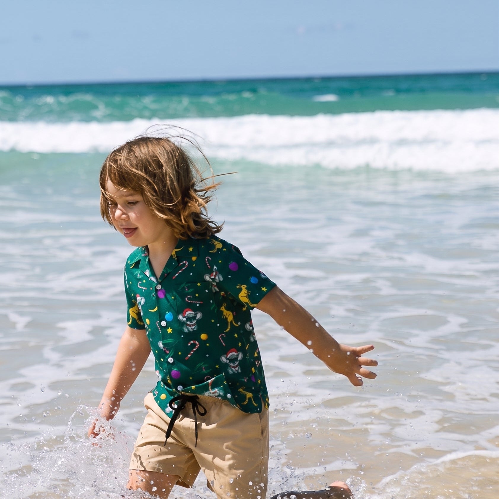 A young boy with light brown hair runs through splashing ocean water on a beach, wearing a dark green short-sleeve button-up shirt with a colorful cartoon print and beige shorts.