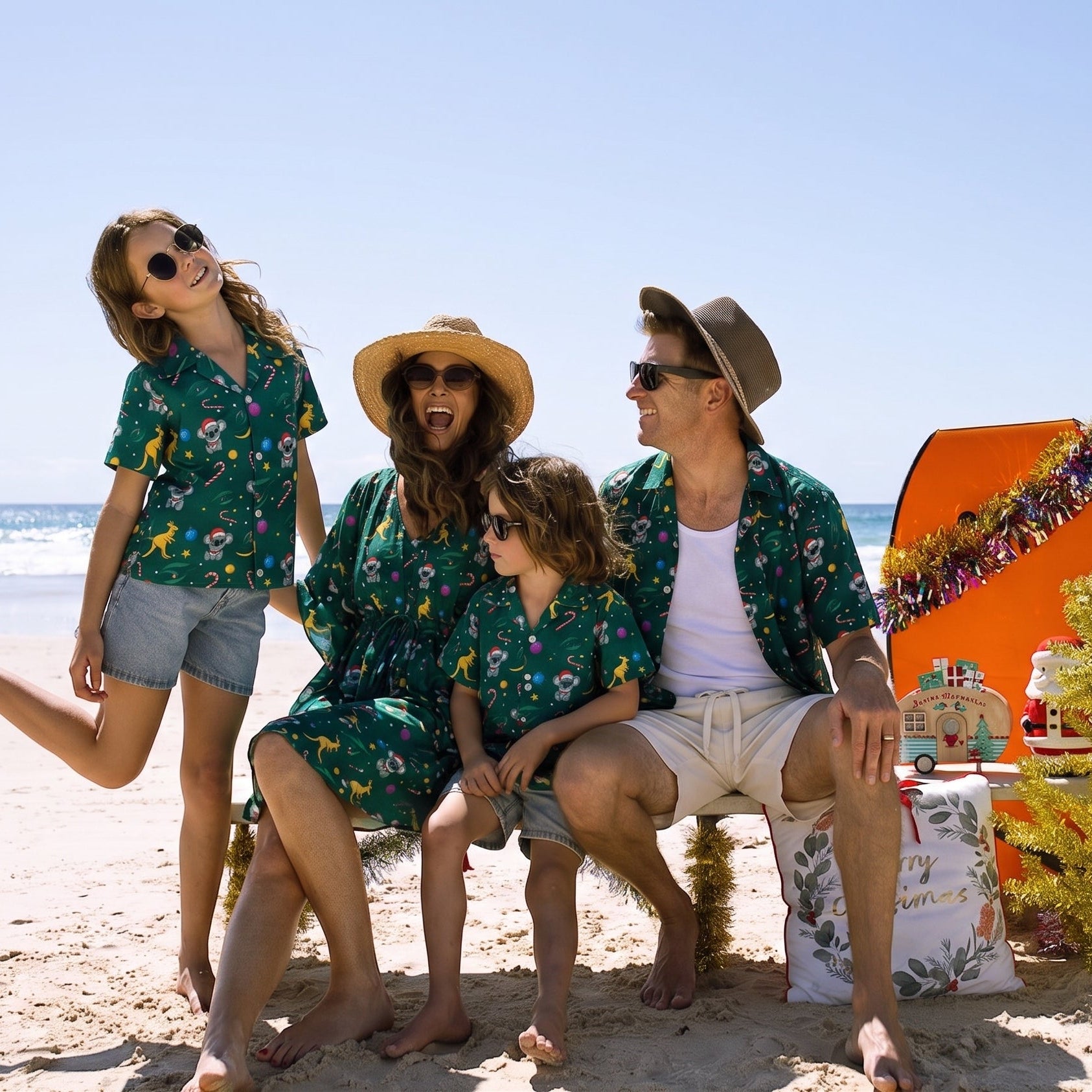 A family of four wearing matching green tropical floral shirts and shorts stands on a sandy beach next to an orange surfboard decorated with Christmas garlands and a Santa Claus figure.