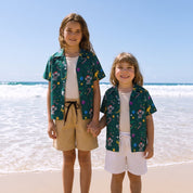 Two children standing on a sandy beach holding hands, wearing matching dark green tropical print short-sleeve button-up shirts over white t-shirts and beige or white shorts.
