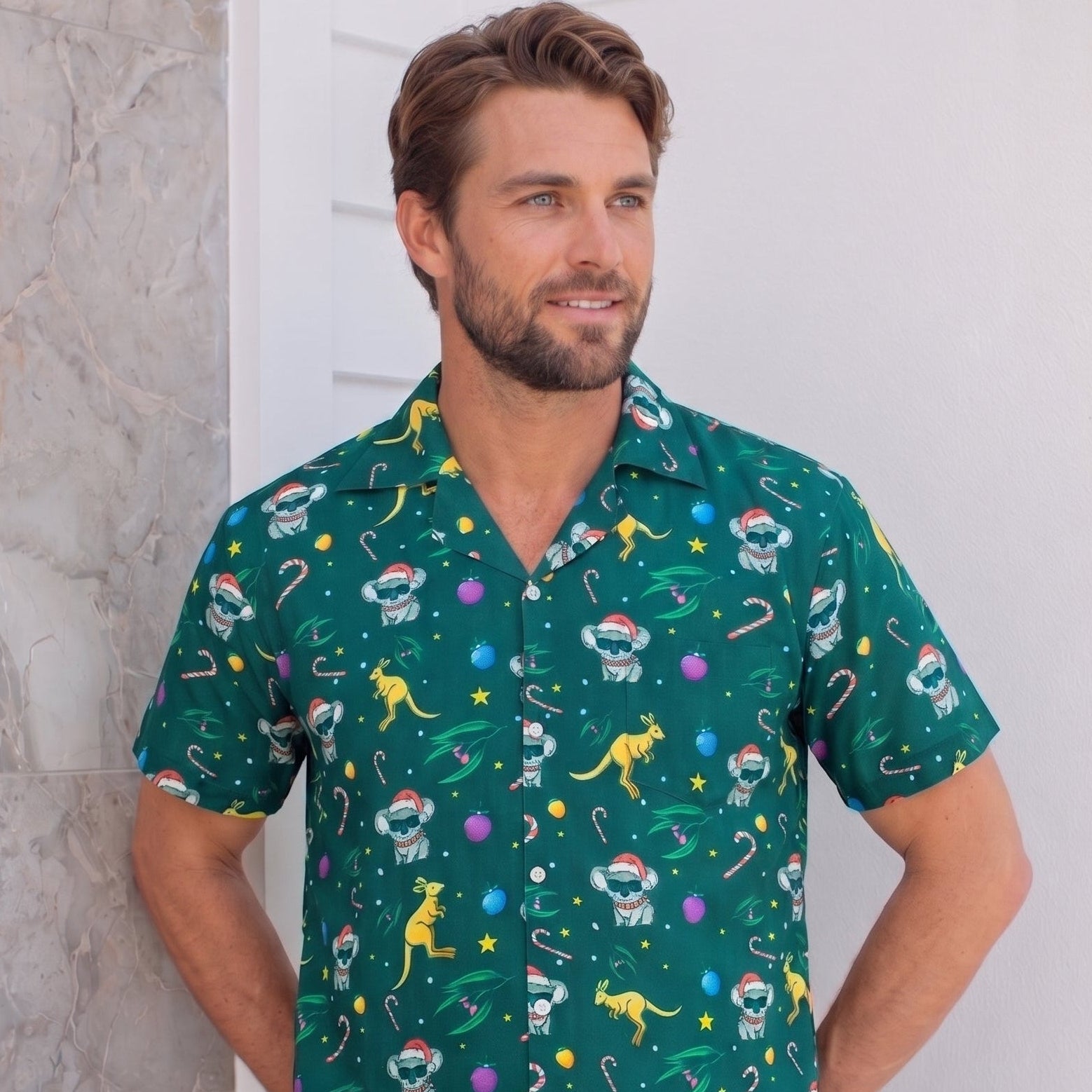 A man wearing a green short-sleeve button-up shirt with a colorful astronaut and dinosaur pattern, paired with black pants, standing against a white wall.