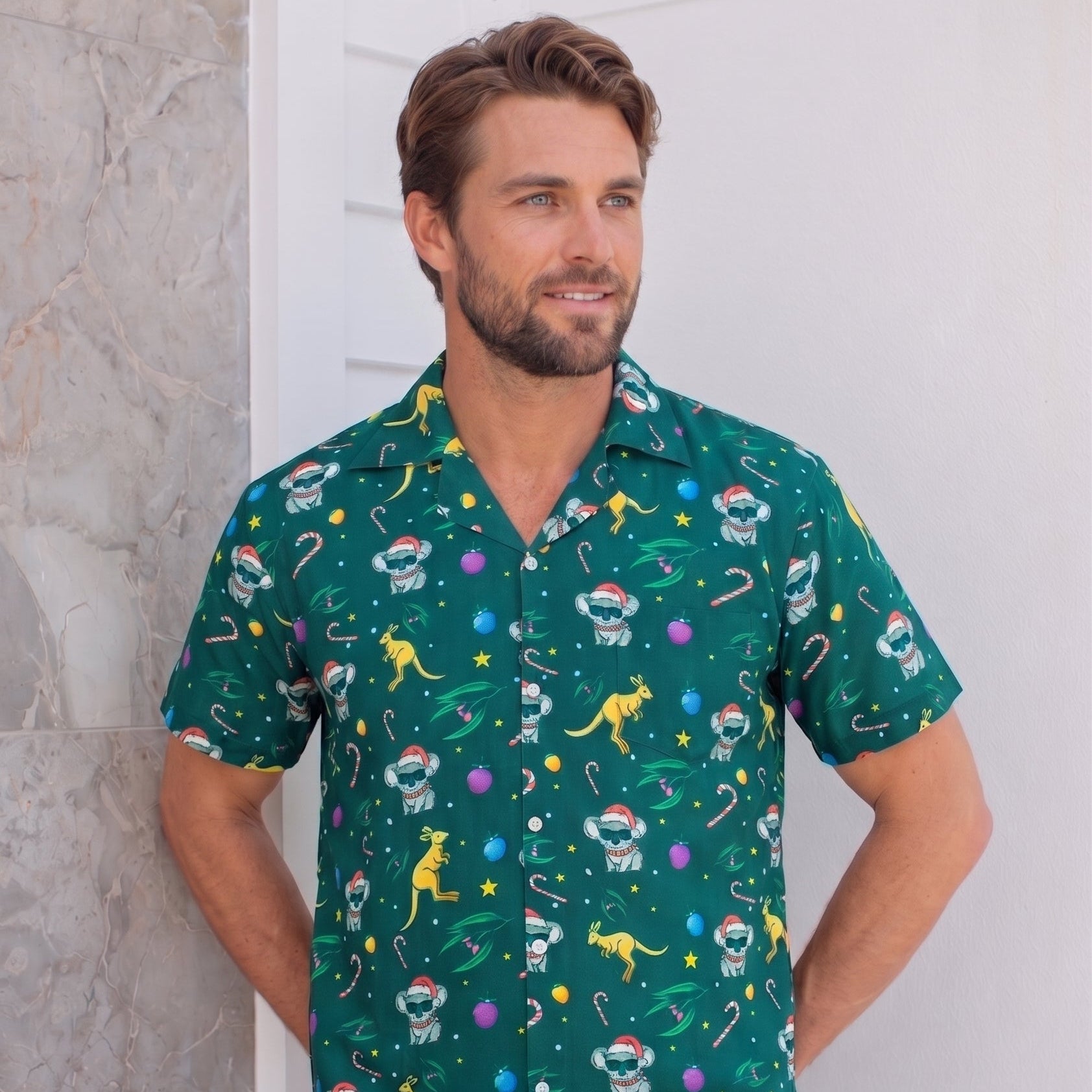 A man wearing a green short-sleeve button-up shirt with a colorful astronaut and dinosaur pattern, paired with black pants, standing against a white wall.
