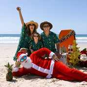 A family of four and a Santa Claus character pose together on a sandy beach. The adults wear matching green tropical print Hawaiian shirts, while the child wears a matching green top.