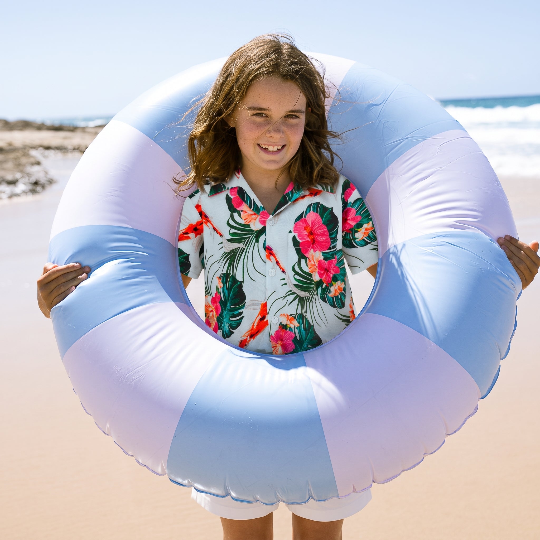 A smiling girl stands on a sandy beach holding a large light blue and white inflatable swim ring. She is wearing a white short-sleeve button-up shirt with a tropical floral print featuring pink flowers and green leaves.