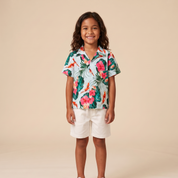A young child stands against a warm beige studio background wearing a short-sleeve tropical print button-up shirt with white shorts and white sneakers.