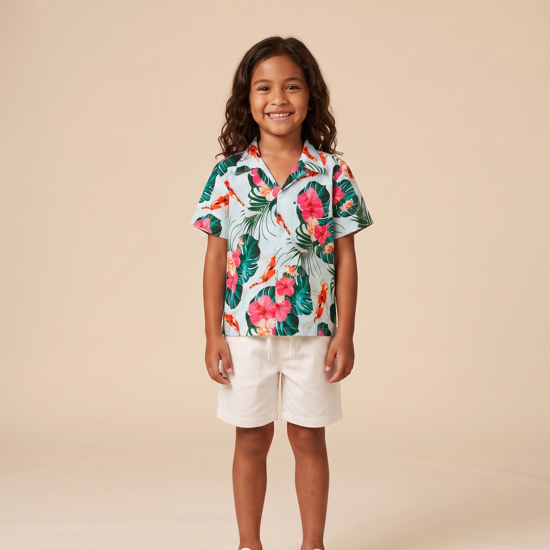 A young child stands against a warm beige studio background wearing a short-sleeve tropical print button-up shirt with white shorts and white sneakers.