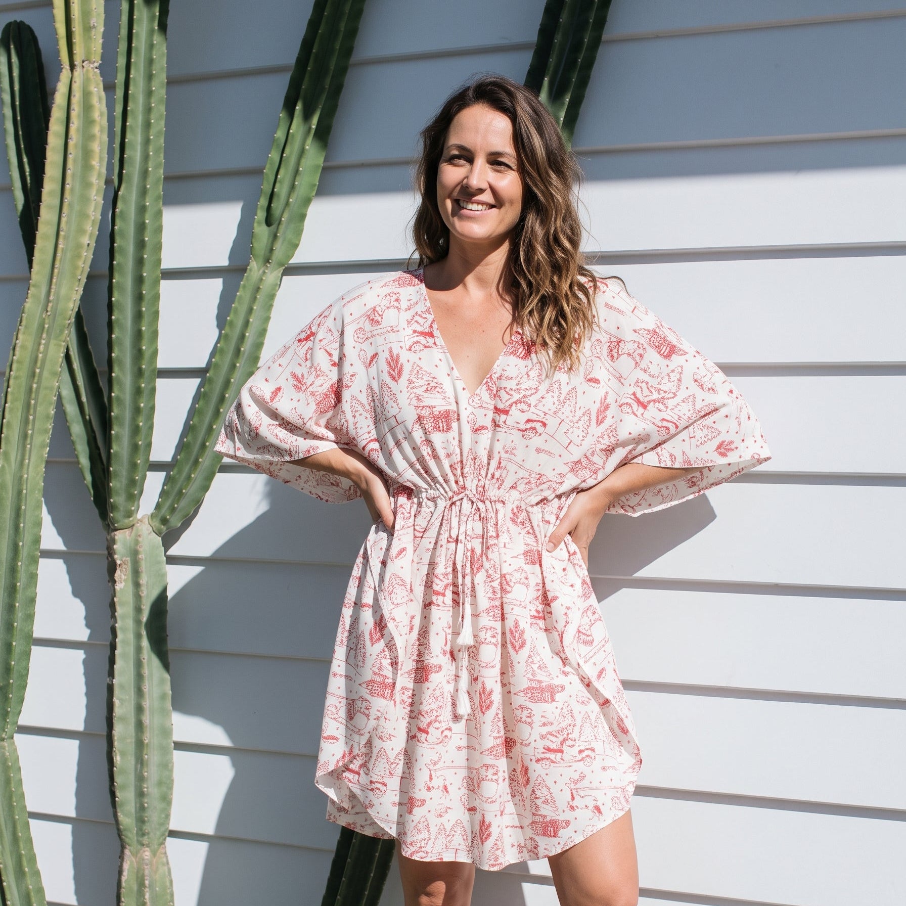 A woman wearing a light pink and white floral print mini dress with wide sleeves, standing outdoors against a white siding wall next to a tall cactus.