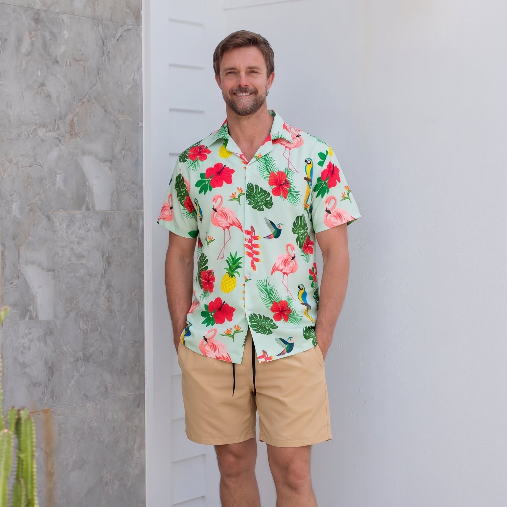 A man wearing a short-sleeve Hawaiian-style camp shirt with a tropical floral and flamingo print, paired with khaki shorts and black slide sandals, standing in a modern indoor setting with a white wall and textured gray stone column.
