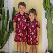 Two children standing barefoot in front of a yellow door, wearing matching short-sleeve pajama sets with a red base and a Santa Claus and holiday print. Tall cacti are visible on both sides of the door.