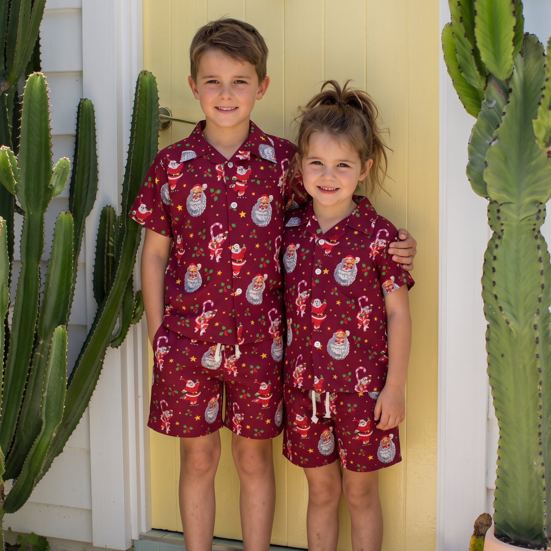 Two children standing barefoot in front of a yellow door, wearing matching short-sleeve pajama sets with a red base and a Santa Claus and holiday print. Tall cacti are visible on both sides of the door.