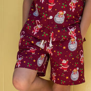 A young boy stands barefoot in front of a yellow door, wearing a matching short-sleeve pajama set with a red base and a festive Santa Claus snowman print. The scene is framed by tall potted cacti on both sides.