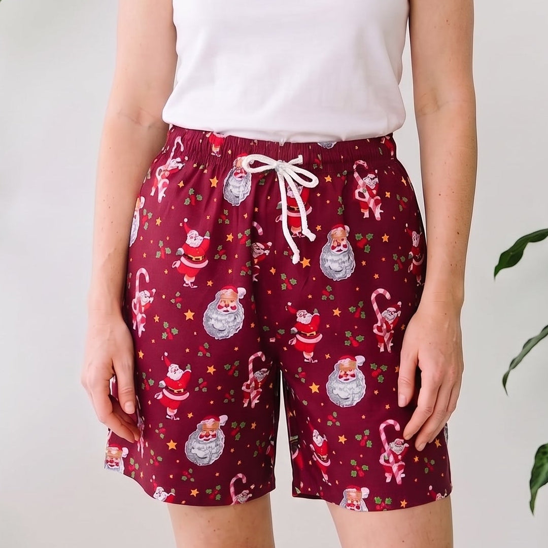 A woman stands indoors wearing a white sleeveless tank top and burgundy holiday print shorts with a white drawstring. Large green potted plants are visible on both sides against a white wall.